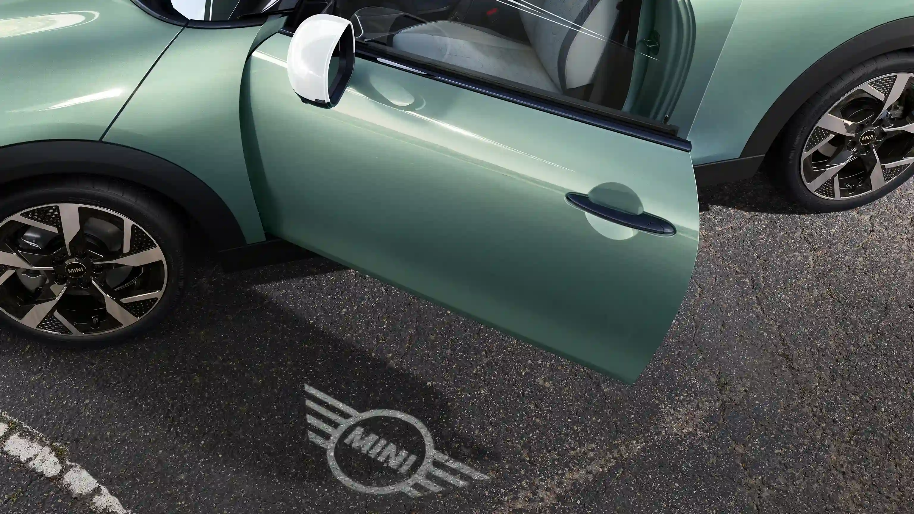 MINI Cooper eksterjero vaizdas su atidarytomis durelėmis ir ant žemės projektuojamu MINI logotipo pasveikinimo apšvietimu
