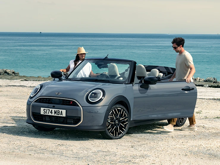 Paplūdimyje stovinčio MINI Cooper kabrioleto pilkos Copper Grey spalvos priekinis vaizdas iš kairės su šalia stovinčiais dviem žmonėmis.