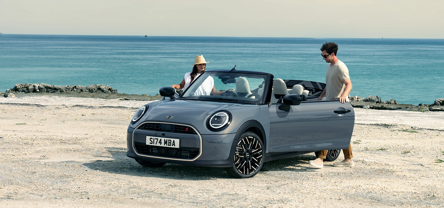 Paplūdimyje stovinčio MINI Cooper kabrioleto pilkos Copper Grey spalvos priekinis vaizdas iš kairės su šalia stovinčiais dviem žmonėmis.