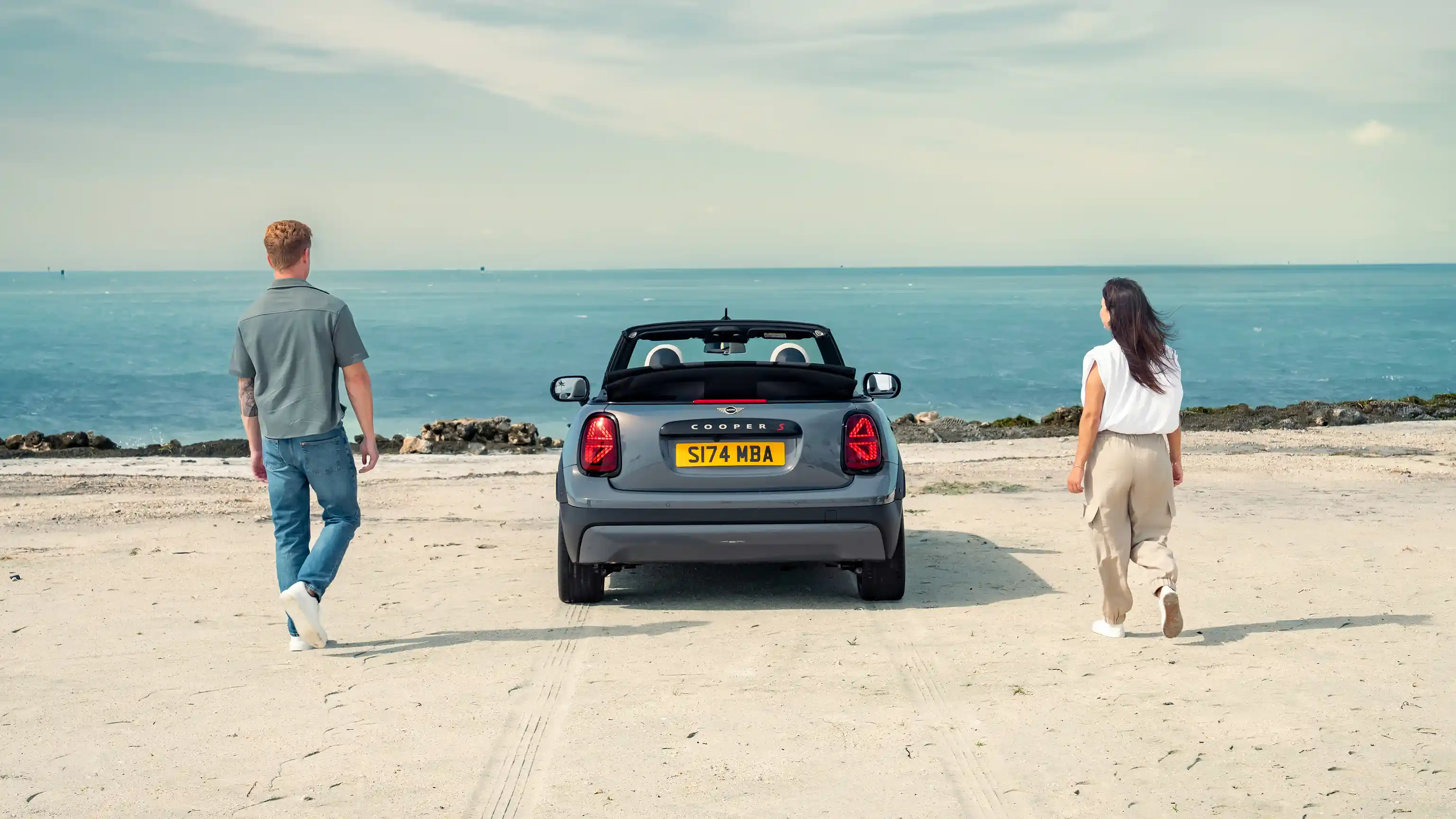 Paplūdimyje stovinčio MINI Cooper kabrioleto vaizdas iš galo su šalia stovinčiais dviem žmonėmis