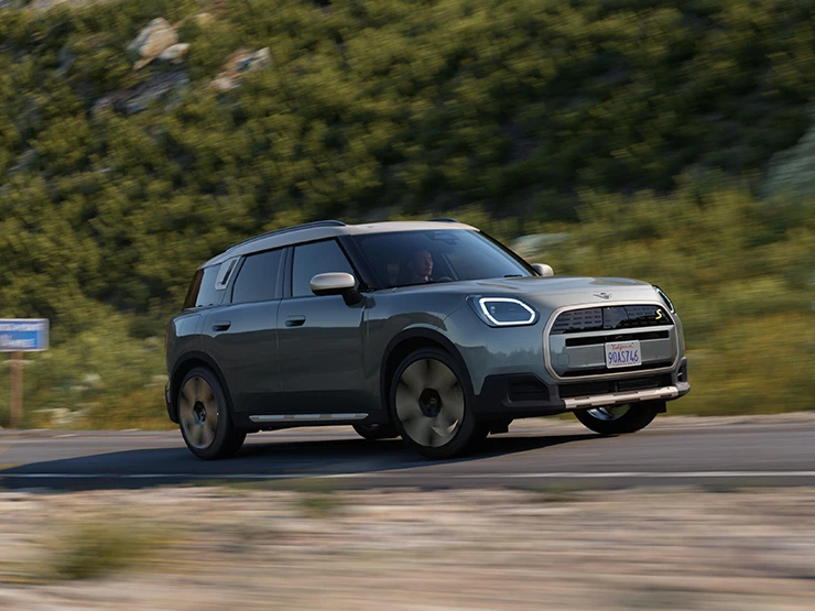 Važiuojančio bekele žalios Smokey Green spalvos visiškai elektrinio MINI Countryman vaizdas iš šono su pilkos spalvos Vibrant Silver stogu