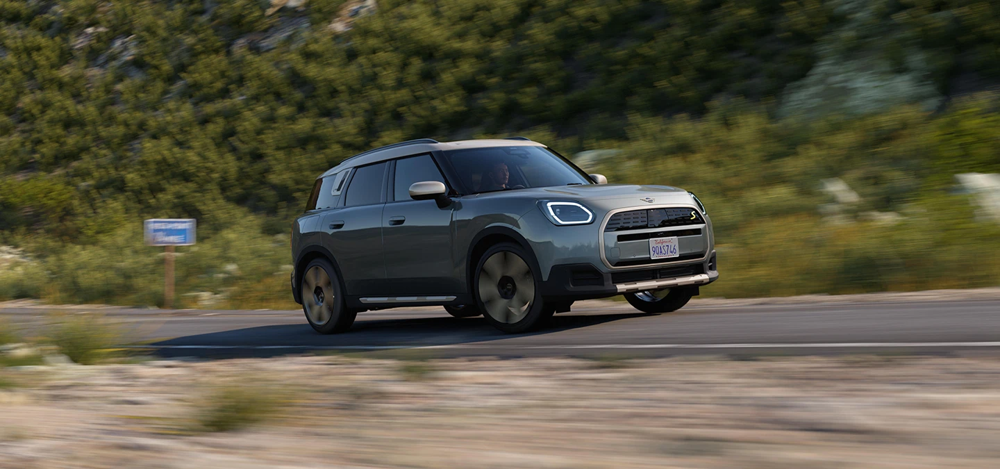 Važiuojančio bekele žalios Smokey Green spalvos visiškai elektrinio MINI Countryman vaizdas iš šono su pilkos spalvos Vibrant Silver stogu
