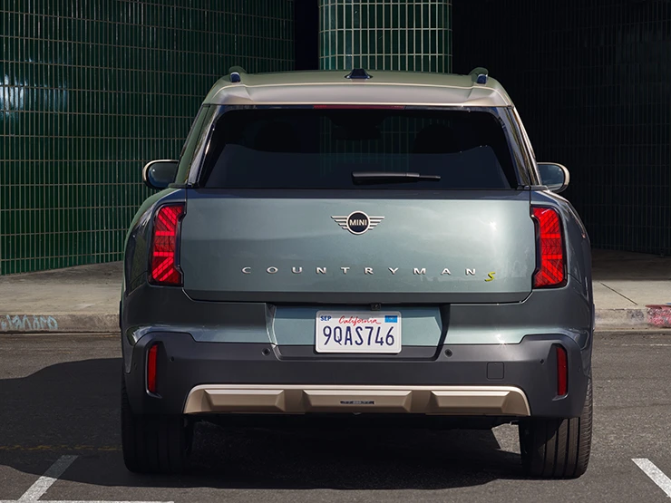 Žalios Smokey Green spalvos visiškai elektrinio MINI Countryman vaizdas iš galo parkinge su pilkos spalvos Vibrant Silver stogu.