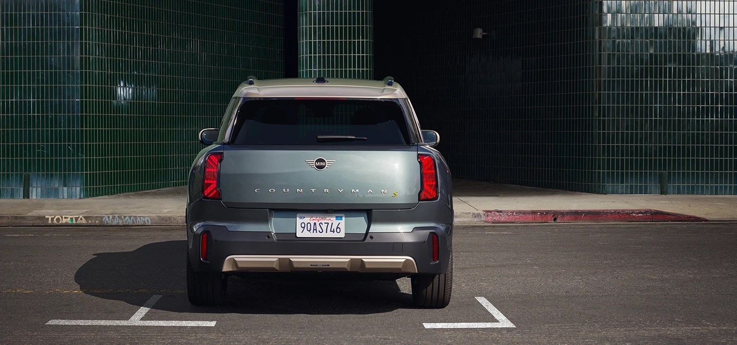 Žalios Smokey Green spalvos visiškai elektrinio MINI Countryman vaizdas iš galo parkinge su pilkos spalvos Vibrant Silver stogu.