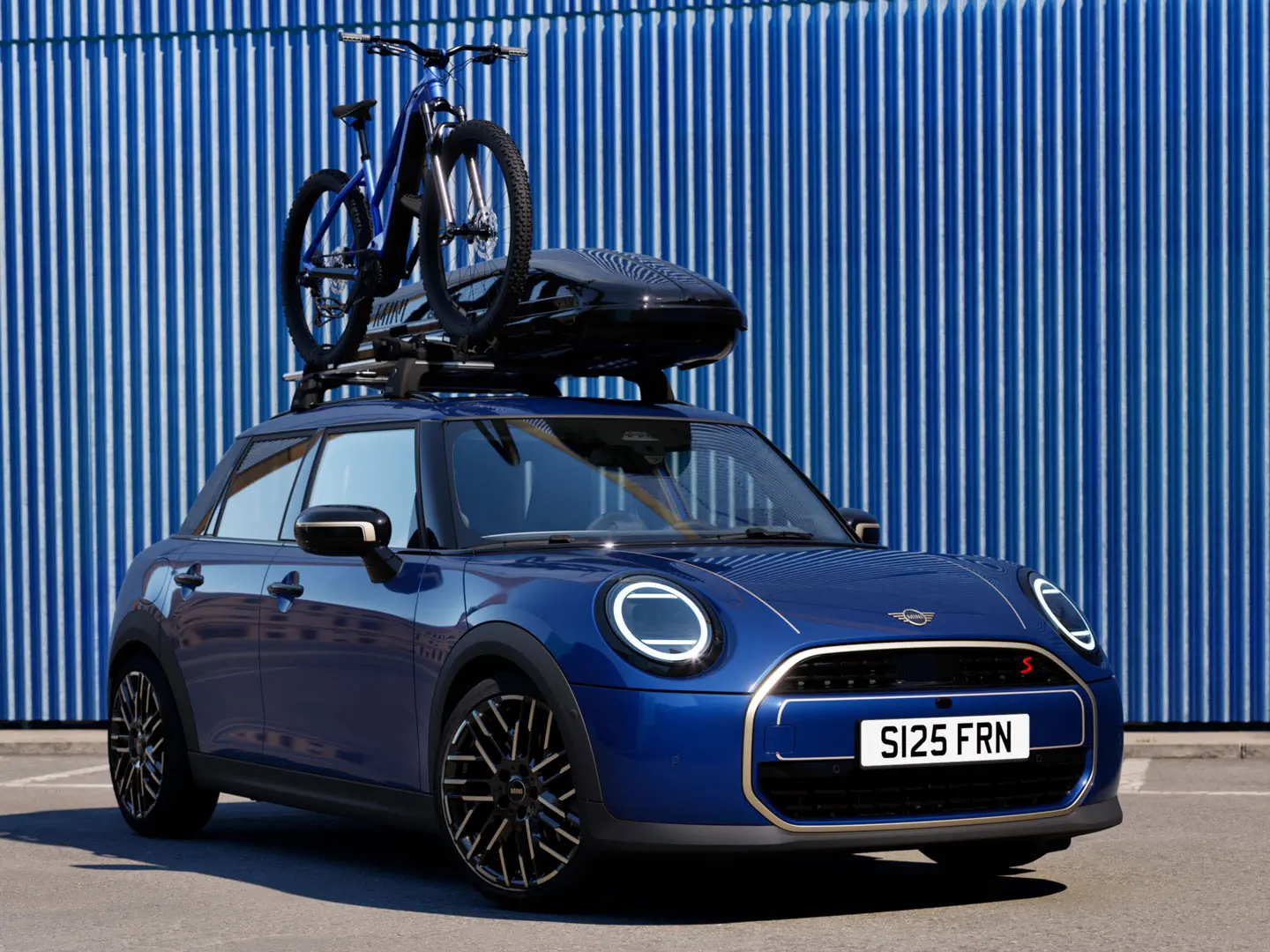 MINI Aceman mėlynos Indigo Sunset Blue spalvos vaizdas iš priekio su MINI ant stogo tvirtinamu dviračių laikikliu, ant kurio pritvirtintas kalnų dviratis ir MINI stogo bagažinė 320.