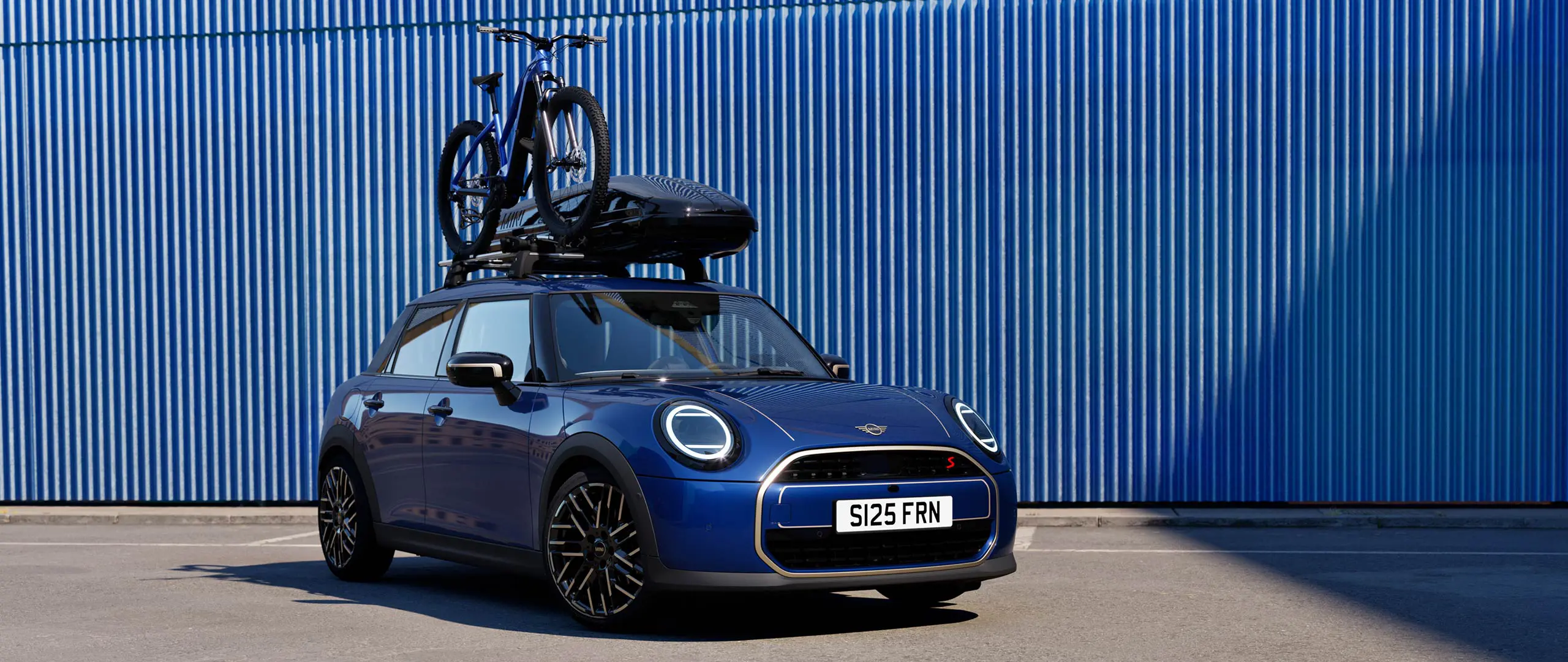 MINI Aceman mėlynos Indigo Sunset Blue spalvos vaizdas iš priekio su MINI ant stogo tvirtinamu dviračių laikikliu, ant kurio pritvirtintas kalnų dviratis ir MINI stogo bagažinė 320.