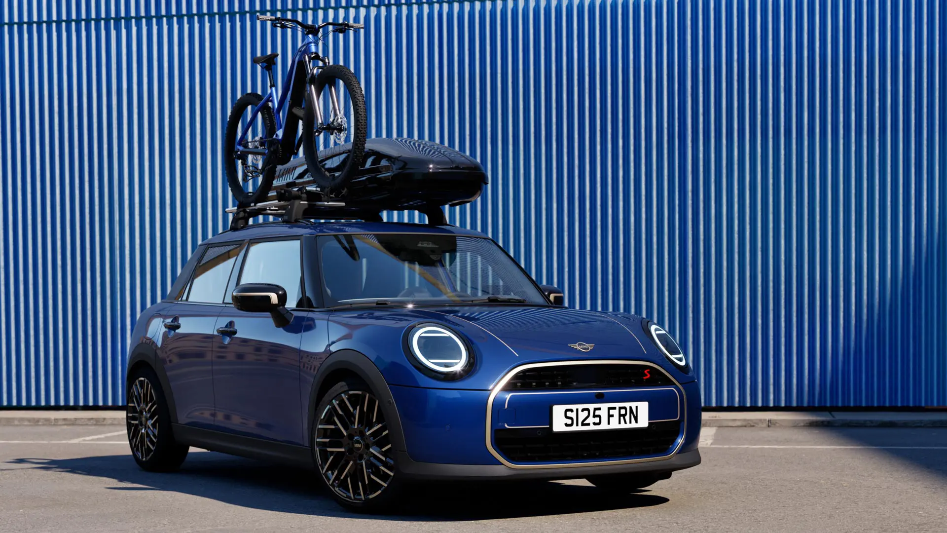 MINI Aceman mėlynos Indigo Sunset Blue spalvos vaizdas iš priekio su MINI ant stogo tvirtinamu dviračių laikikliu, ant kurio pritvirtintas kalnų dviratis ir MINI stogo bagažinė 320.