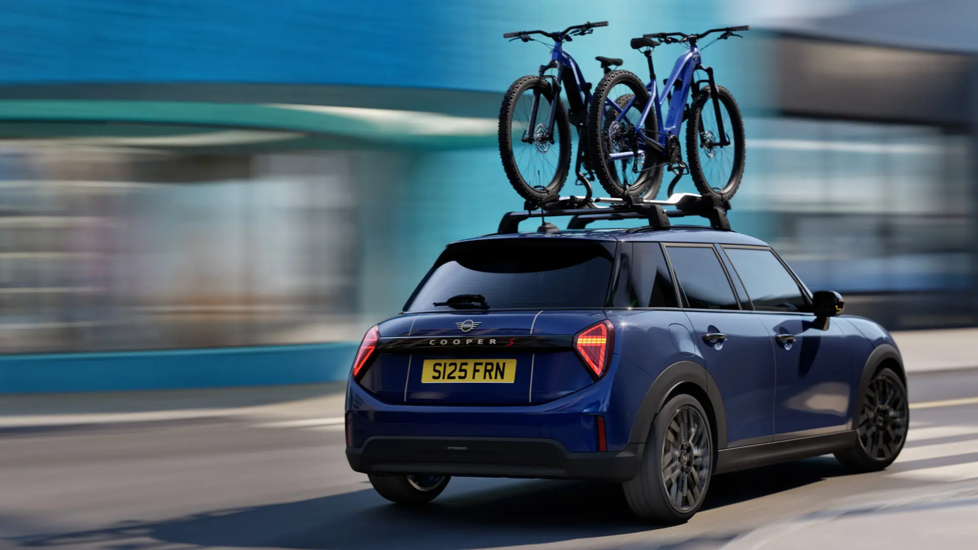 Mėlynos Indigo Sunset Blue spalvos MINI Aceman su MINI ant stogo tvirtinamu dviračių laikikliu, ant kurio transportuojami du kalnų dviračiai, trijų ketvirčių vaizdas iš šono.