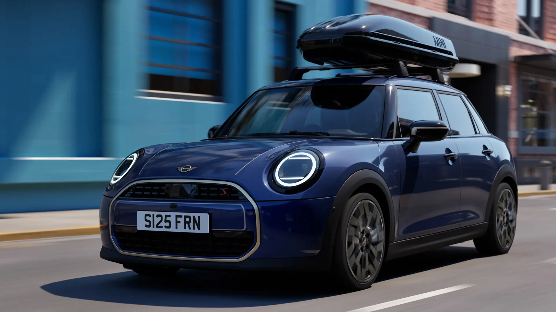 MINI Aceman mėlynos Indigo Sunset Blue spalvos trijų ketvirčių vaizdas iš priekio su MINI stogo skersiniais, prie kurių pritvirtinta MINI stogo bagažinė 320